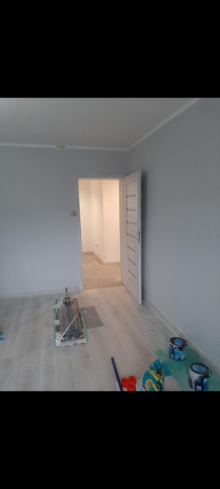 Wnętrze w trakcie malowania na szaro, widoczny otwarty pokój, drzwi z białymi panelami, na podłodze narzędzia do malowania, pędzle, kuwety, wiaderka z farbą i maszyna do cięcia płytek.