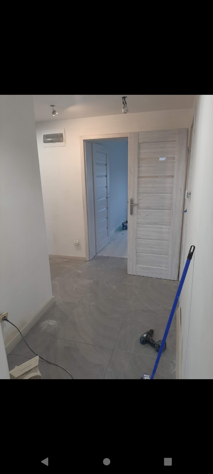 Wnętrze mieszkania w trakcie remontu: szare płytki podłogowe, świeżo pomalowane białe ściany, dwoje białych drzwi z przeszkleniami oraz narzędzia - wiertarka i kij do malowania.