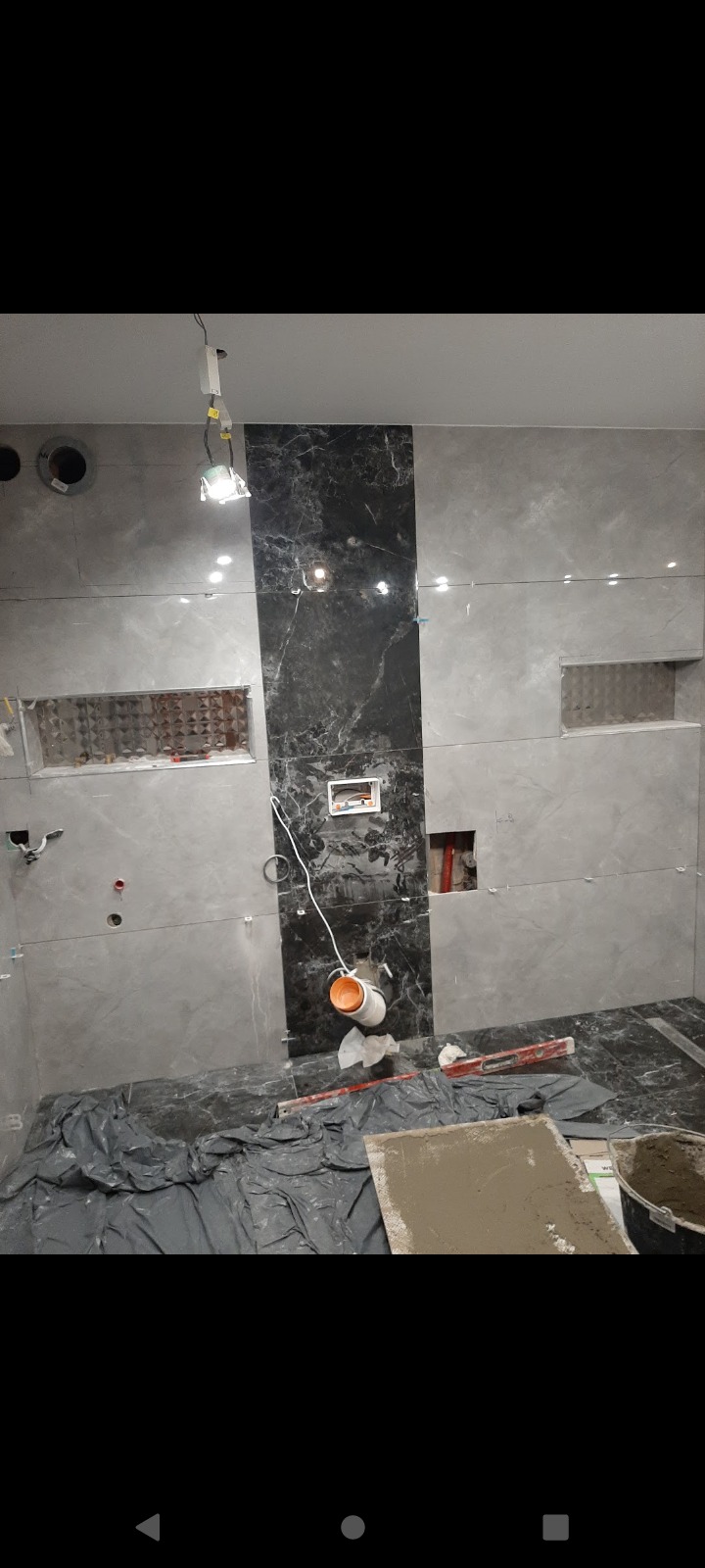 Łazienka w trakcie remontu: szare i czarne płytki marmurowe na ścianach, wycięte wnęki, poziomica, wiadro z zaprawą, folia ochronna na podłodze, widoczne instalacje.