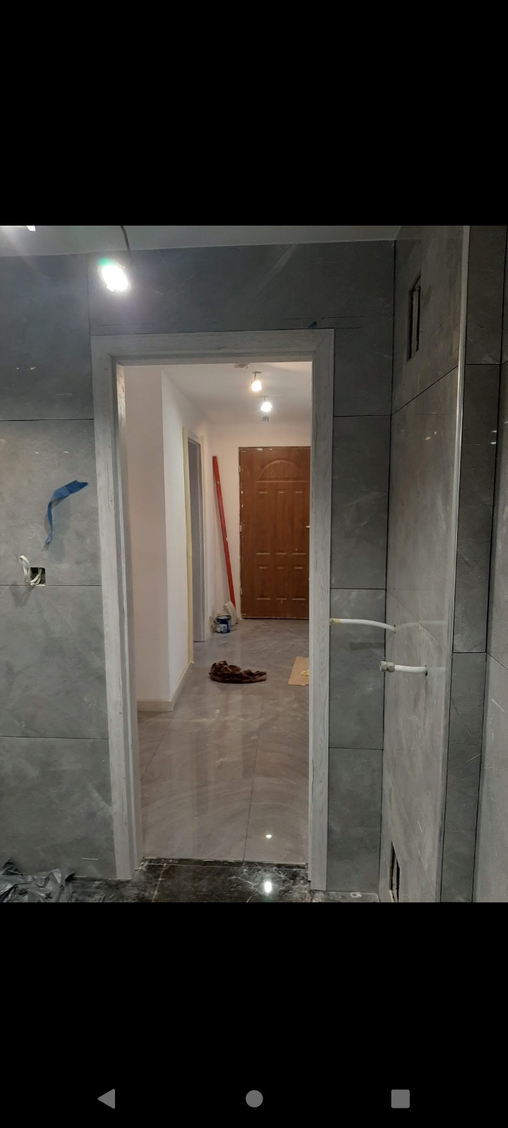 Wnętrze łazienki w trakcie remontu: szare płytki imitujące marmur na ścianach i podłodze, widoczne rury i niedokończone wykończenie ościeżnicy drzwiowej, w tle widoczny korytarz z brązowymi drzwiami.