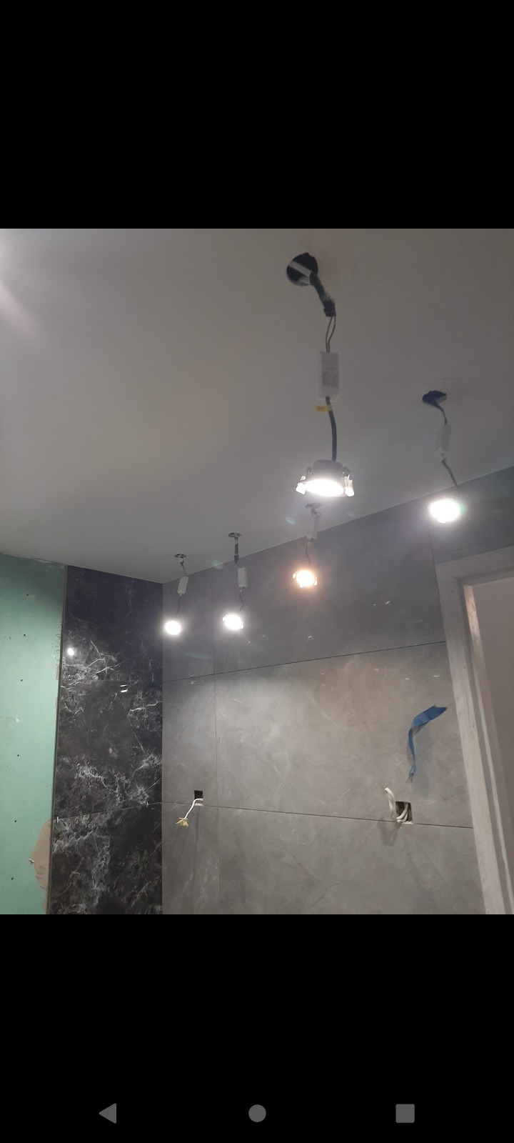 Wnętrze łazienki w trakcie remontu: ściany częściowo wyłożone płytkami imitującymi marmur, częściowo szara gładź, widoczne przewody elektryczne i punkty świetlne w suficie przed instalacją opraw...