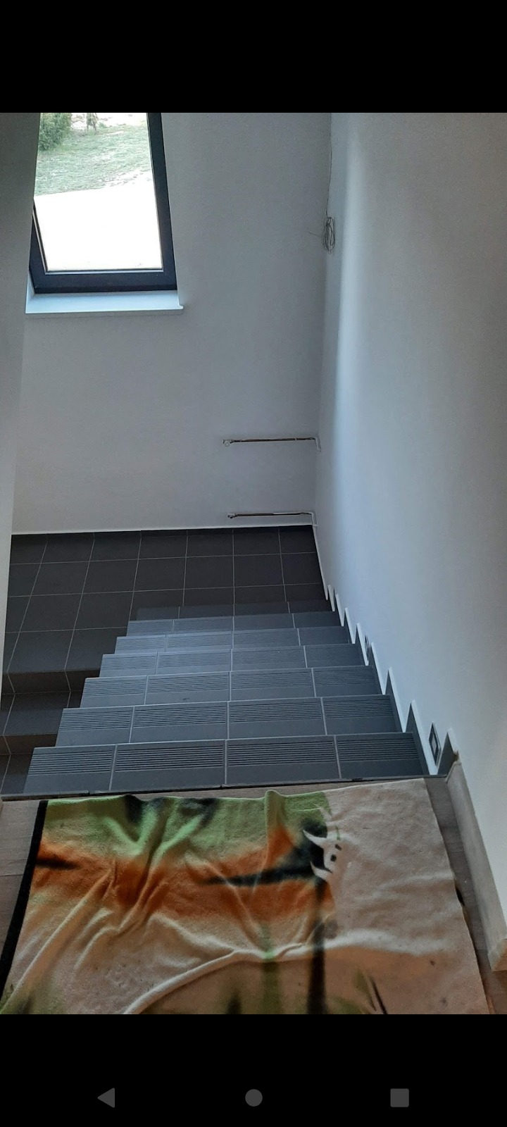 Wnętrze domu w trakcie wykańczania: widok z góry na schody z ciemnoszarej terakoty, białe ściany z widocznymi instalacjami, okno dachowe i koc z wzorem jelenia na podłodze.