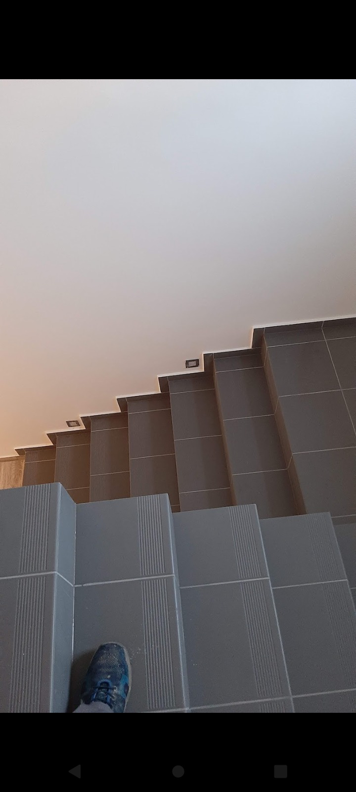 Wnętrze domu: Schody wyłożone szarymi płytkami z delikatnym wzorem, oświetlenie punktowe na ścianie, widok z góry na stopę w niebieskim bucie.