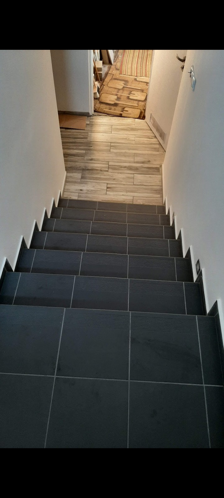 Schody wewnątrz budynku, wyłożone ciemnymi, kwadratowymi płytkami z delikatnym wzorem, prowadzące do pomieszczenia z podłogą z płytek imitujących drewno.