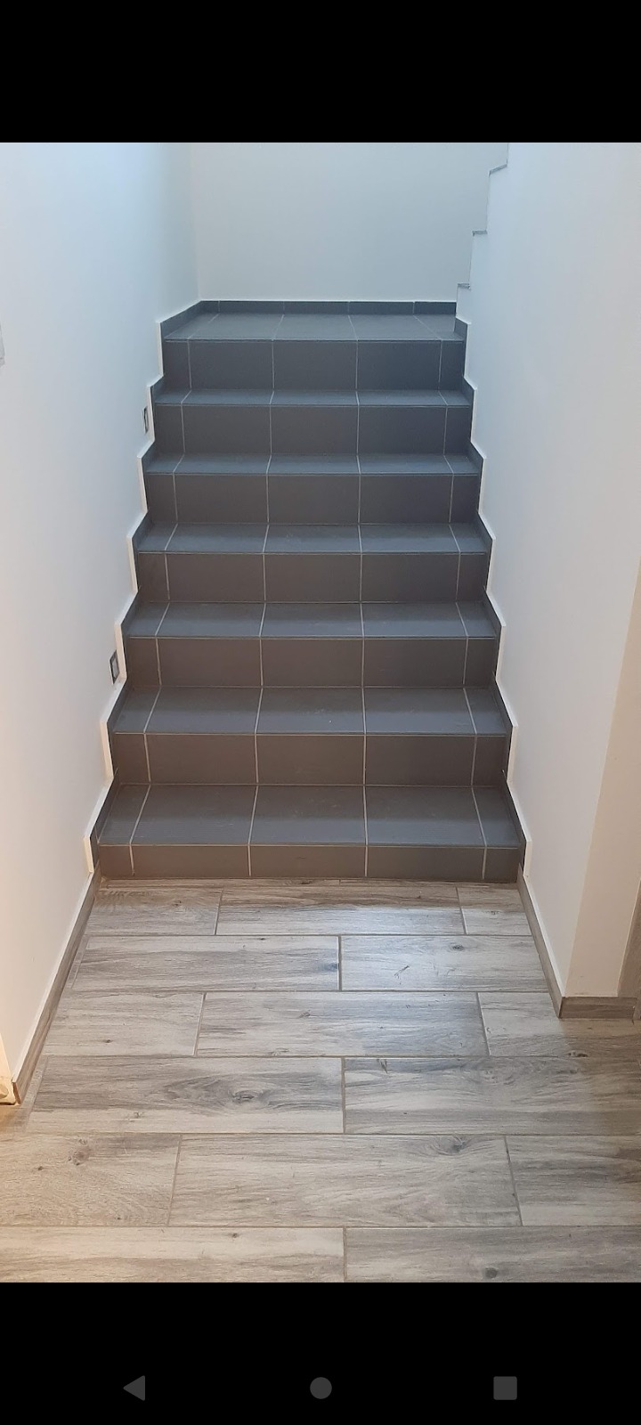 Schody wewnętrzne wyłożone szarymi płytkami ceramicznymi, z oświetleniem punktowym w ścianie, połączone z podłogą imitującą drewno.