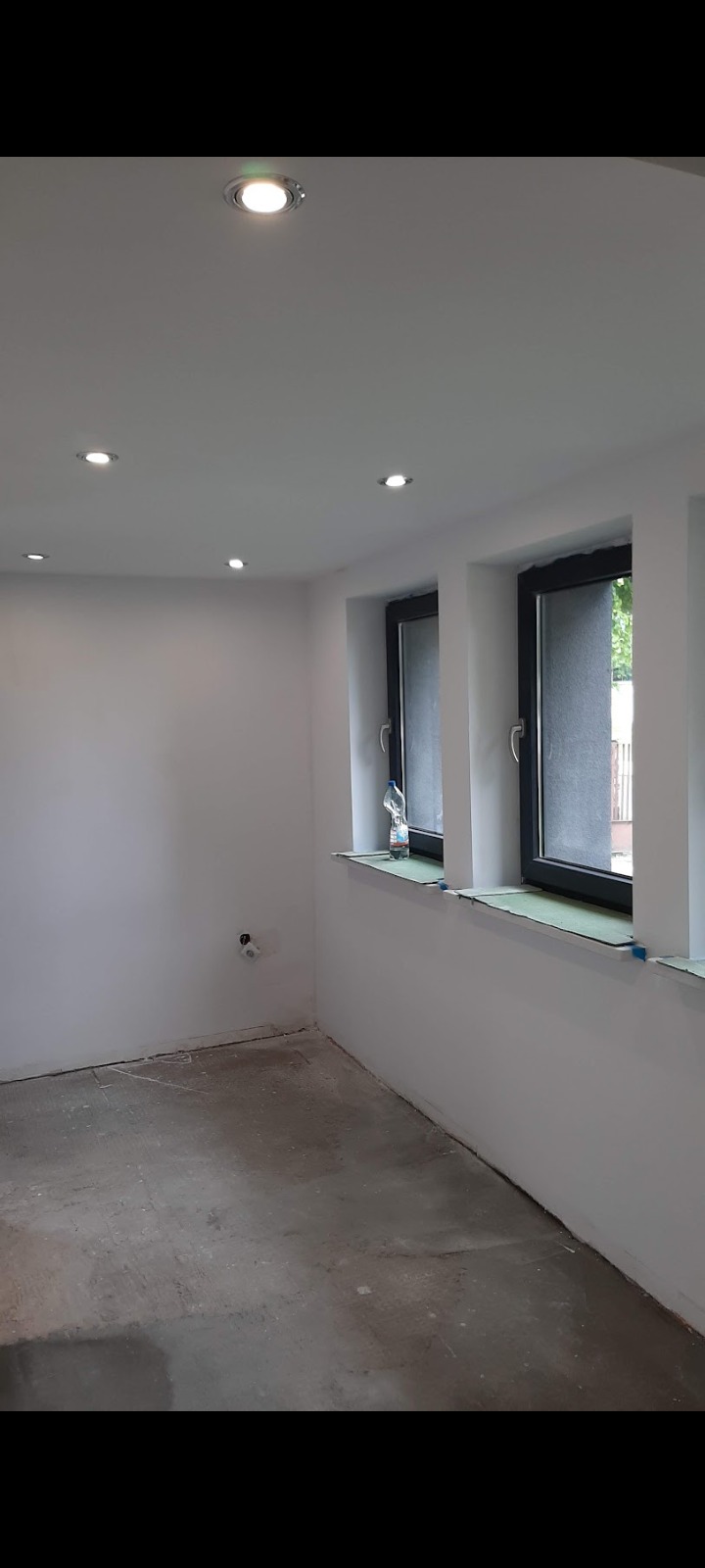 Wnętrze pokoju po remoncie: białe ściany, czarne ramy okienne, szary betonowy podkład podłogowy, sufit z oświetleniem punktowym.