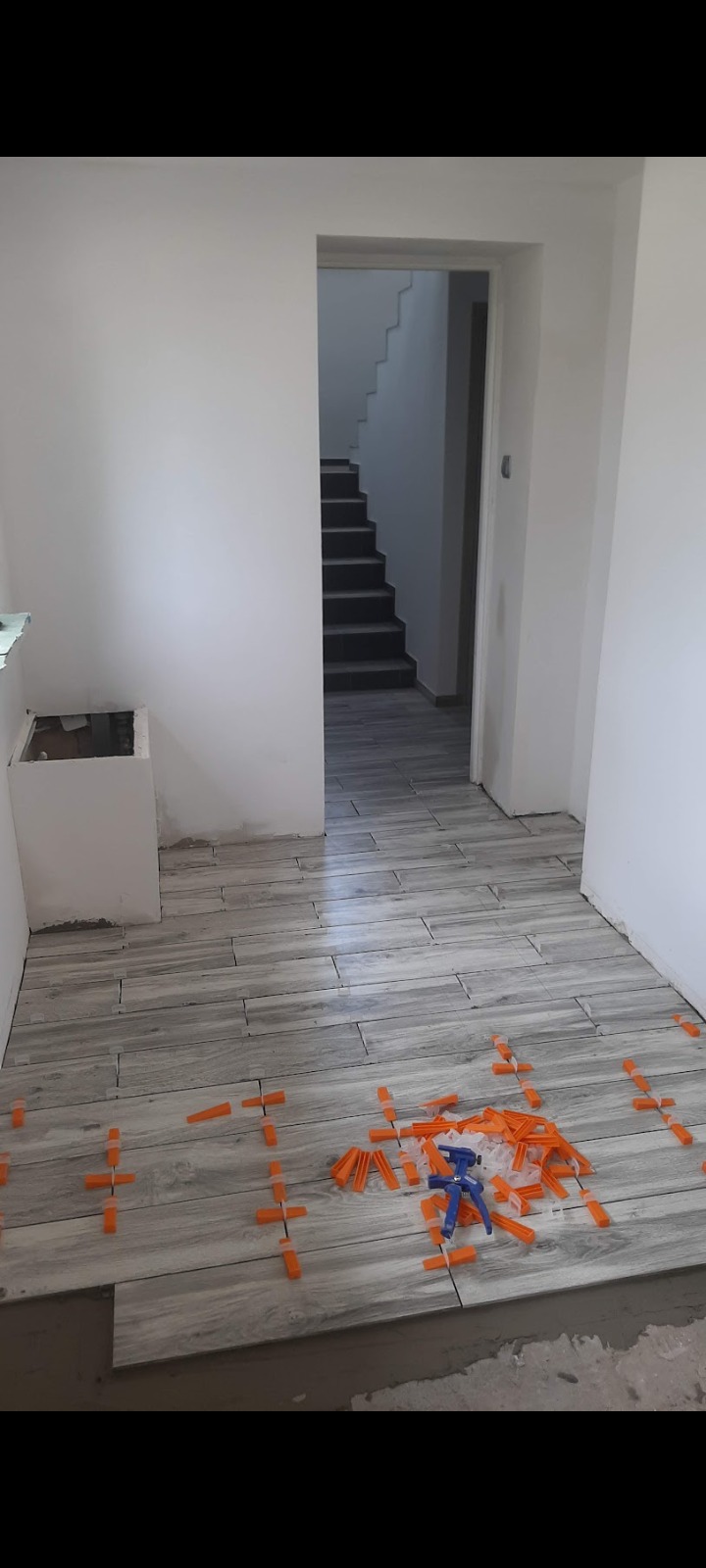 Układanie szarych płytek podłogowych w korytarzu, widoczne pomarańczowe krzyżyki dystansowe i niebieski przyrząd do układania płytek, w tle schody.