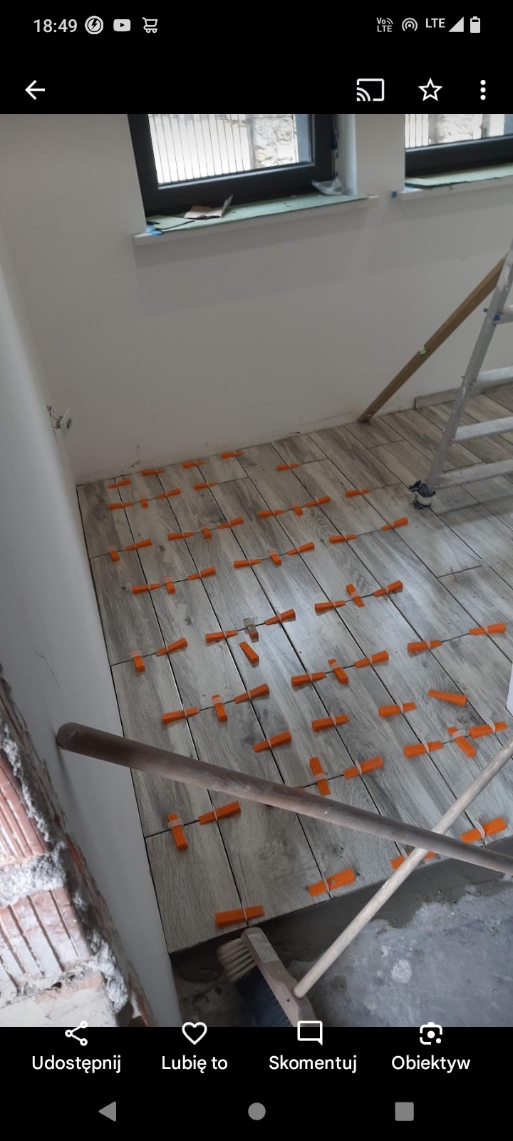 Układanie płytek podłogowych imitujących drewno z użyciem pomarańczowych krzyżyków dystansowych, widoczna drabina i fragment ściany w trakcie wykończenia.