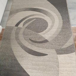 BąbeL - Beżowo-szary dywan z abstrakcyjnym spiralnym wzorem leżący na drewnianej podłodze w rogu pokoju.