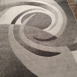 BąbeL - Szaro-beżowy dywan z abstrakcyjnym, spiralnym wzorem, widoczny z góry, z fragmentem drewnianej podłogi w tle.