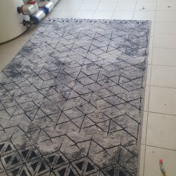 BąbeL - Szary dywan z geometrycznym wzorem, leżący na kafelkach w Ząbkach, obok widoczne rury i akcesoria do czyszczenia.