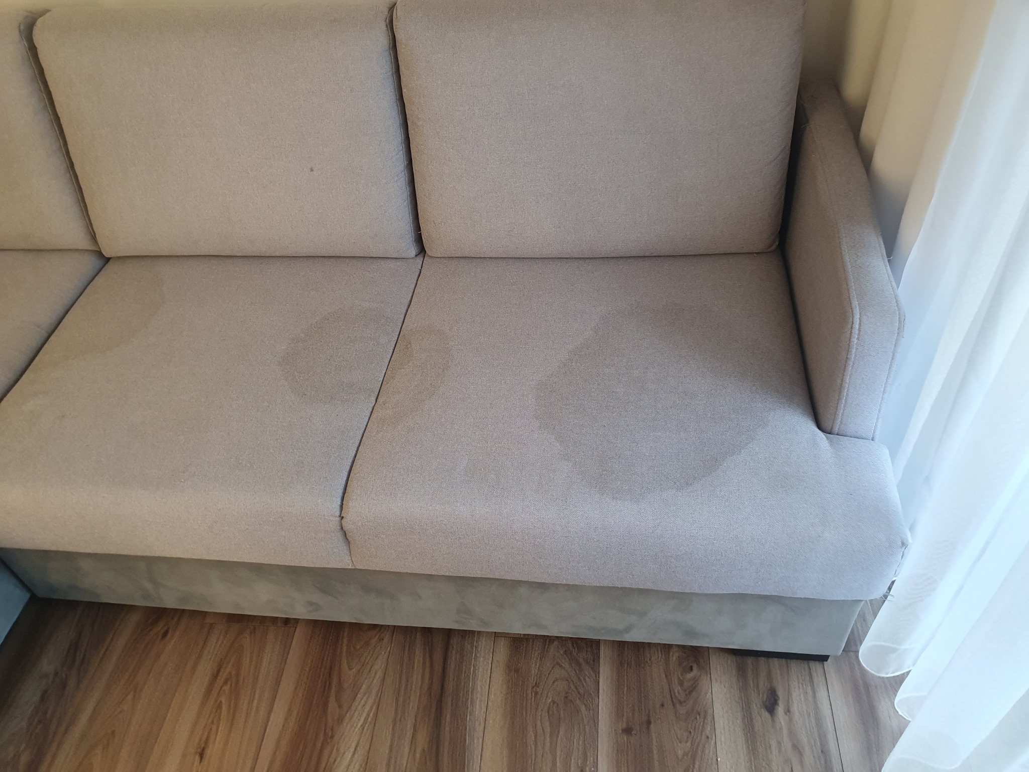 Beżowa kanapa z widocznymi plamami na siedzisku, stojąca na drewnianej podłodze, obok zasłony.