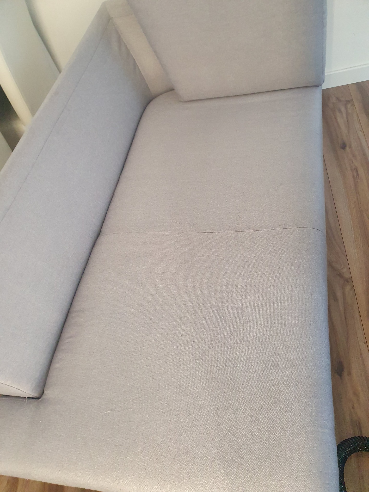 Jasnoszara kanapa z widocznym szwem na siedzisku i fragmentem węża odkurzacza piorącego w prawym dolnym rogu, ukazana z perspektywy górnej.