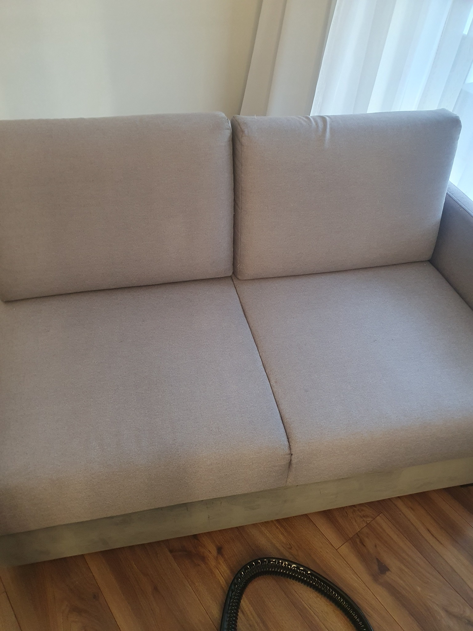 Jasnoszara, dwuosobowa kanapa na drewnianej podłodze, z widoczną końcówką węża od odkurzacza piorącego.