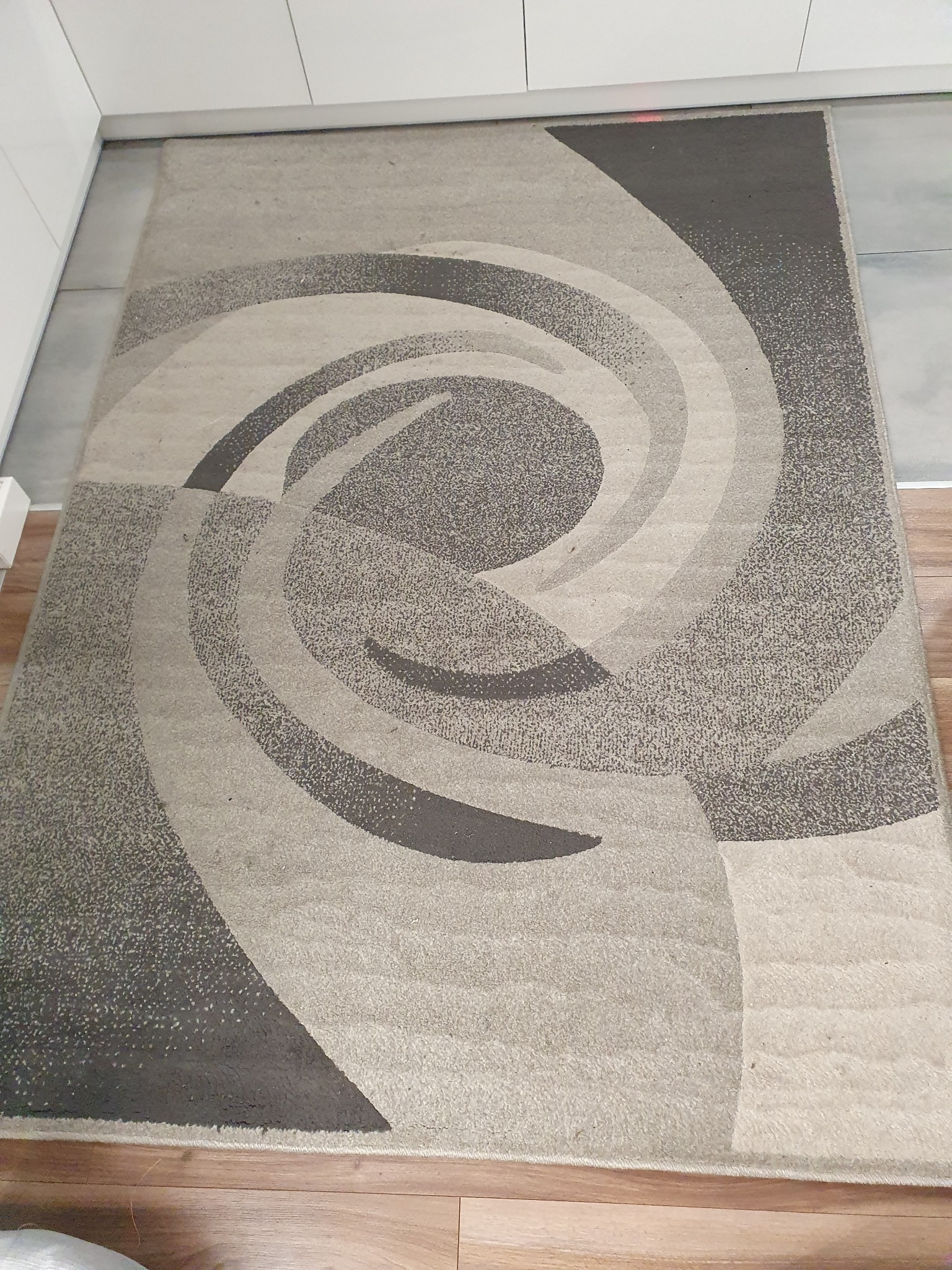 Beżowo-szary dywan z abstrakcyjnym spiralnym wzorem leżący na drewnianej podłodze w rogu pokoju.