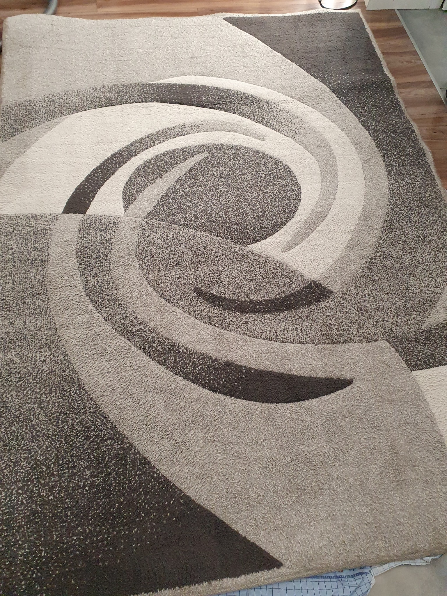 Szaro-beżowy dywan z abstrakcyjnym, spiralnym wzorem, widoczny z góry, z fragmentem drewnianej podłogi w tle.