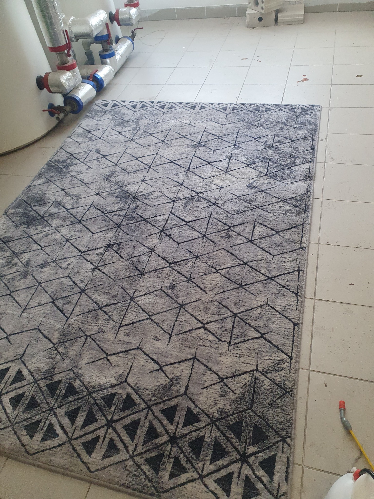 Szary dywan z geometrycznym wzorem, leżący na kafelkach w Ząbkach, obok widoczne rury i akcesoria do czyszczenia.
