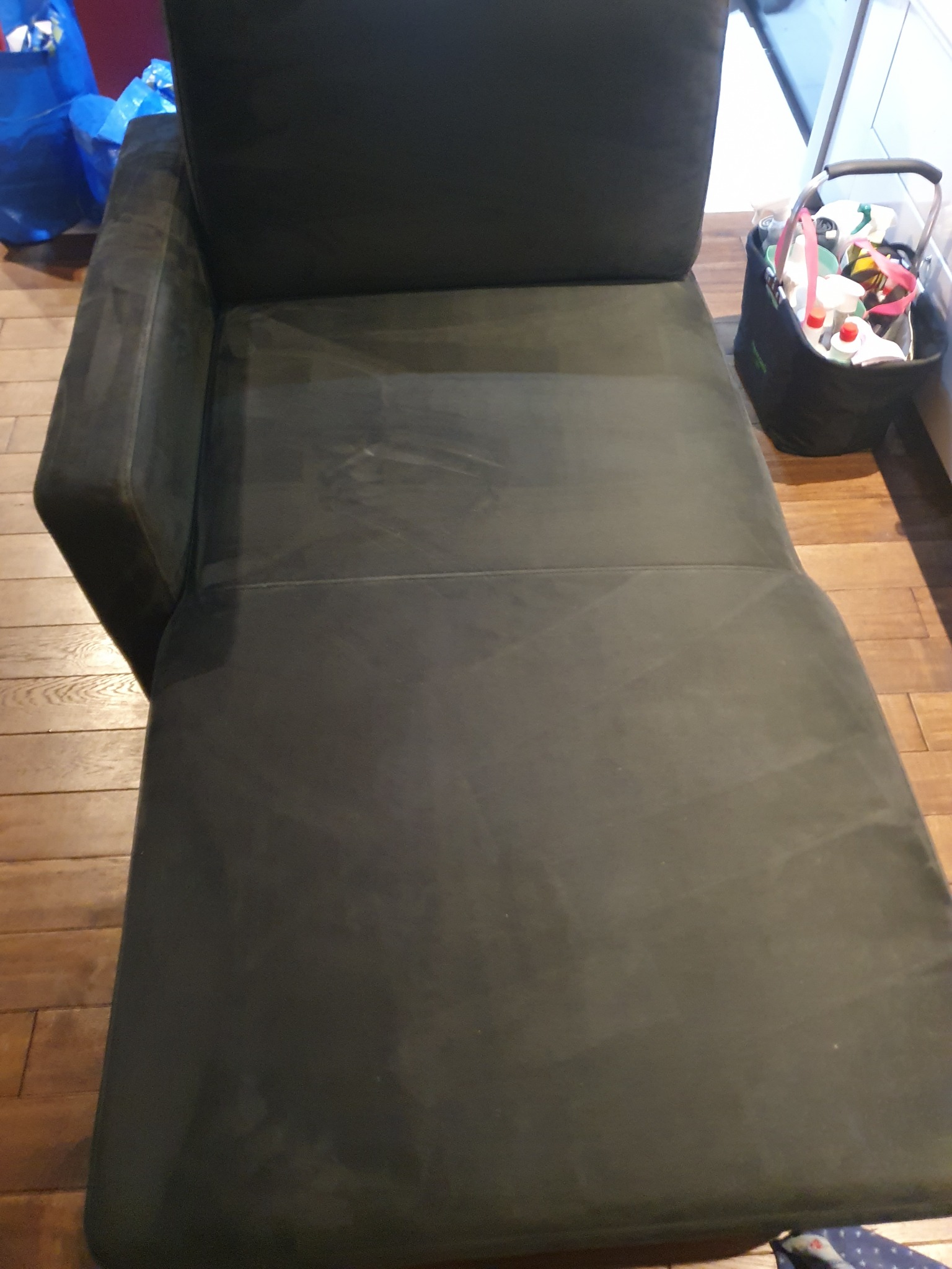 Ciemnoszara leżanka z widocznymi śladami czyszczenia na tapicerce, obok koszyk z detergentami i środkami czystości na drewnianej podłodze.