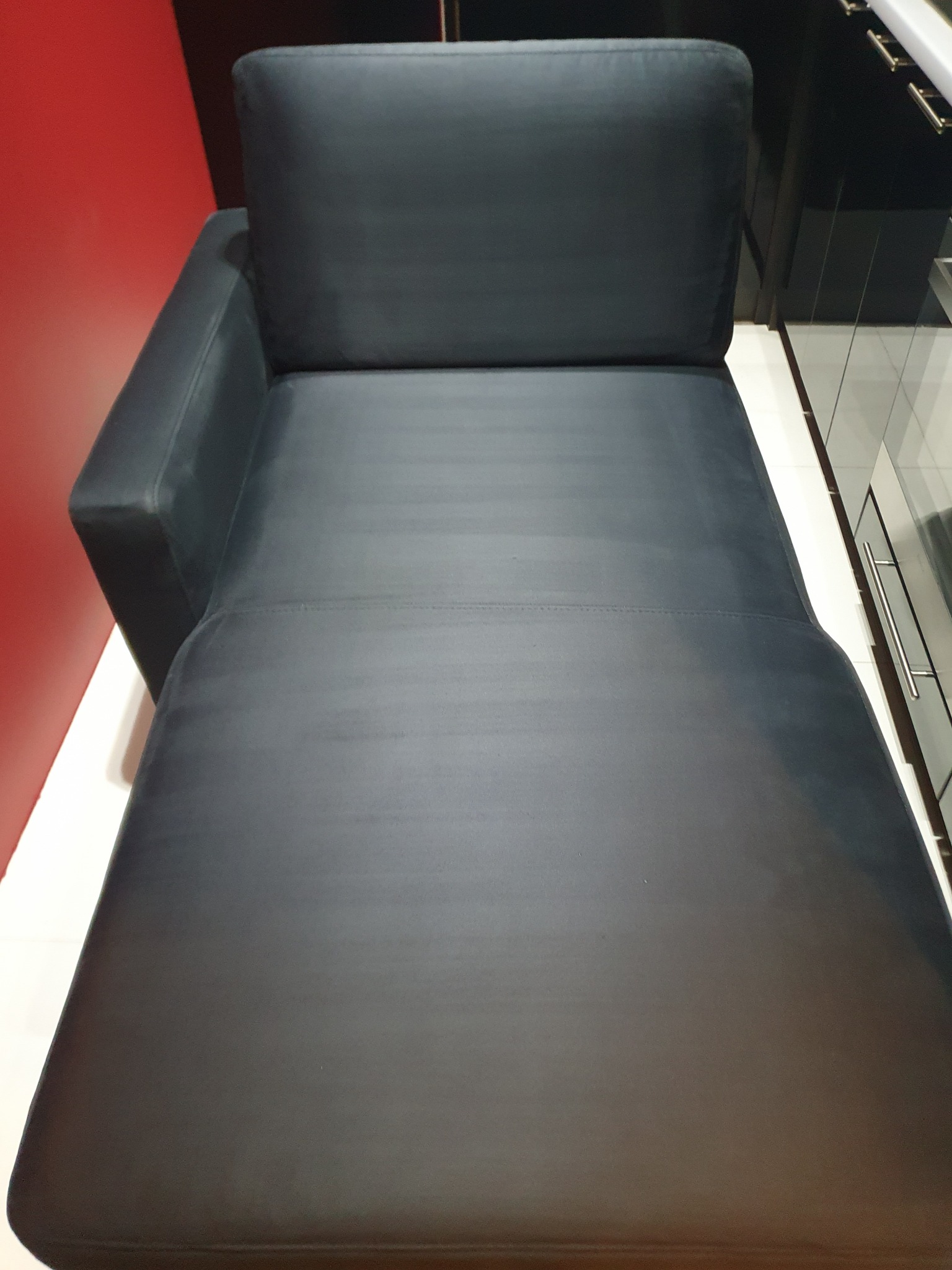 Ciemnoszara, skórzana leżanka z jednym podłokietnikiem na tle czerwonej ściany i czarnych szafek.