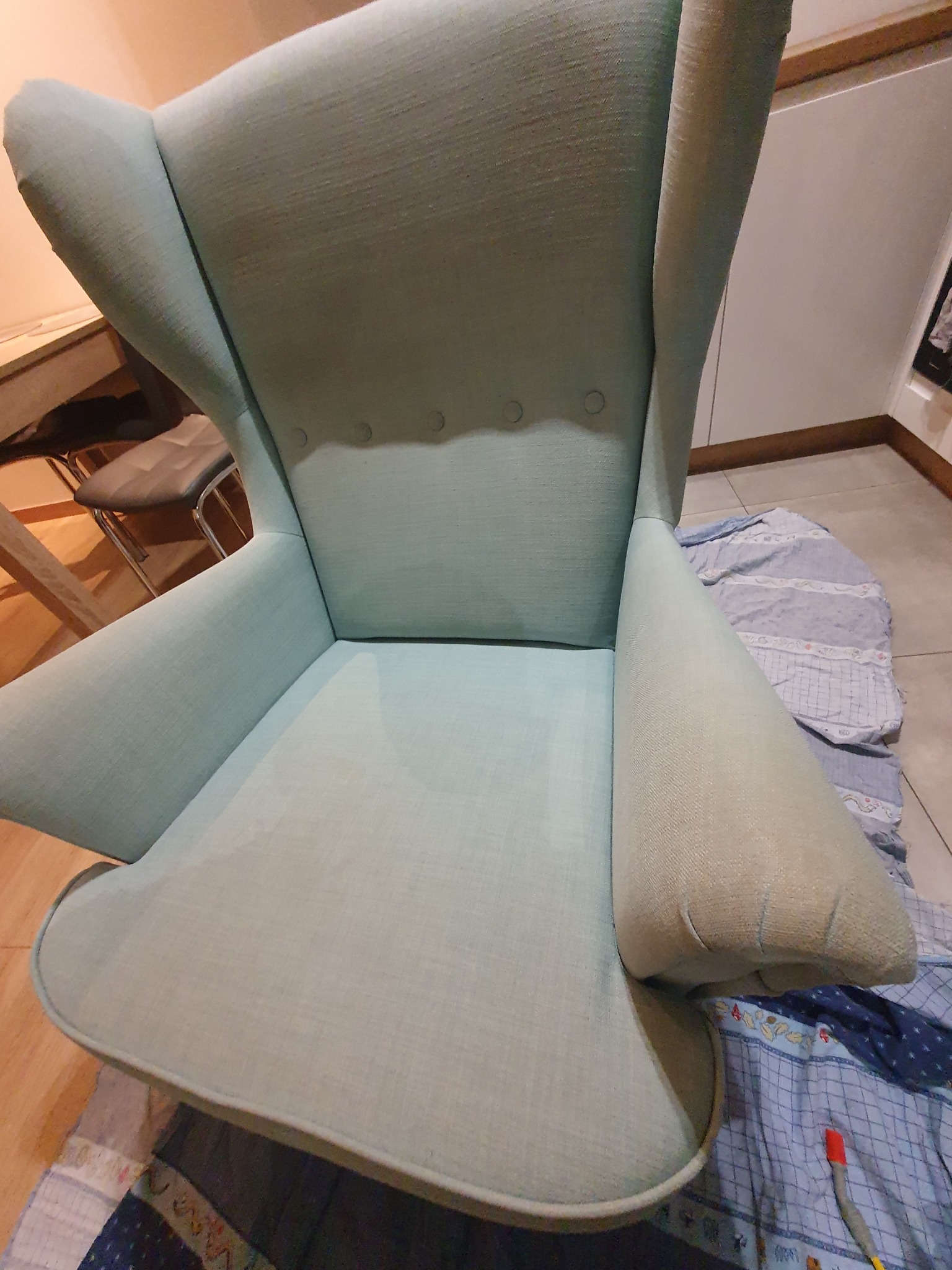 Jasnozielony fotel typu uszak z widoczną strukturą tkaniny, częściowo przykryty wzorzystą narzutą, ukazujący detale tapicerki i ogólny stan mebla.