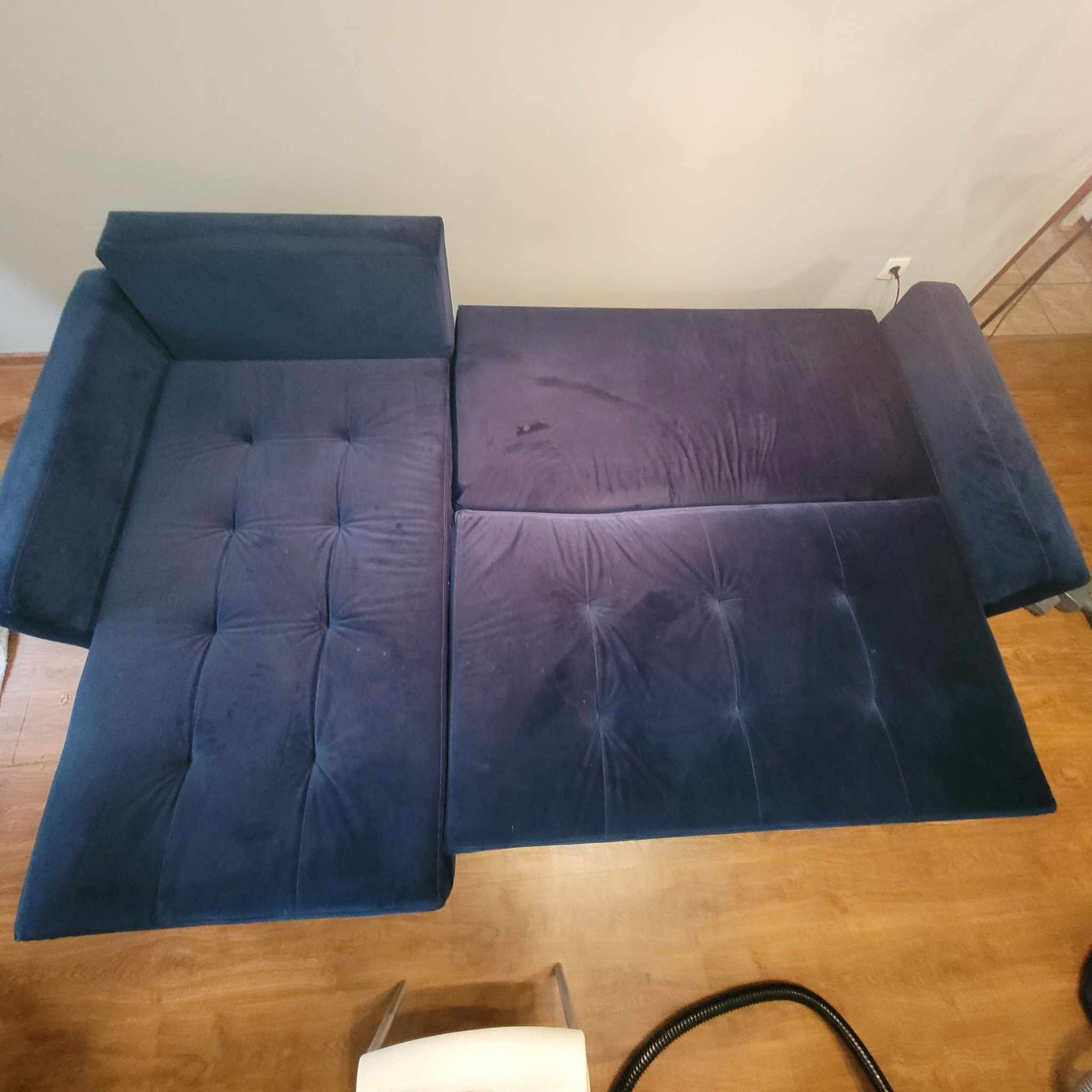 Rozłożona, granatowa kanapa z widocznymi plamami i zaciekami, częściowo czyszczona odkurzaczem piorącym.