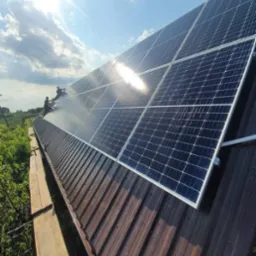 Montaż paneli słonecznych na brązowym dachu blaszanym, dwóch instalatorów pracuje na panelach, widok z dołu na błękitne niebo z chmurami.