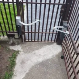 Zewnętrzny widok na brązową bramę skrzydłową z zamontowaną automatyką, widoczne siłowniki i ramię napędowe, świeżo wylany beton wokół słupków.