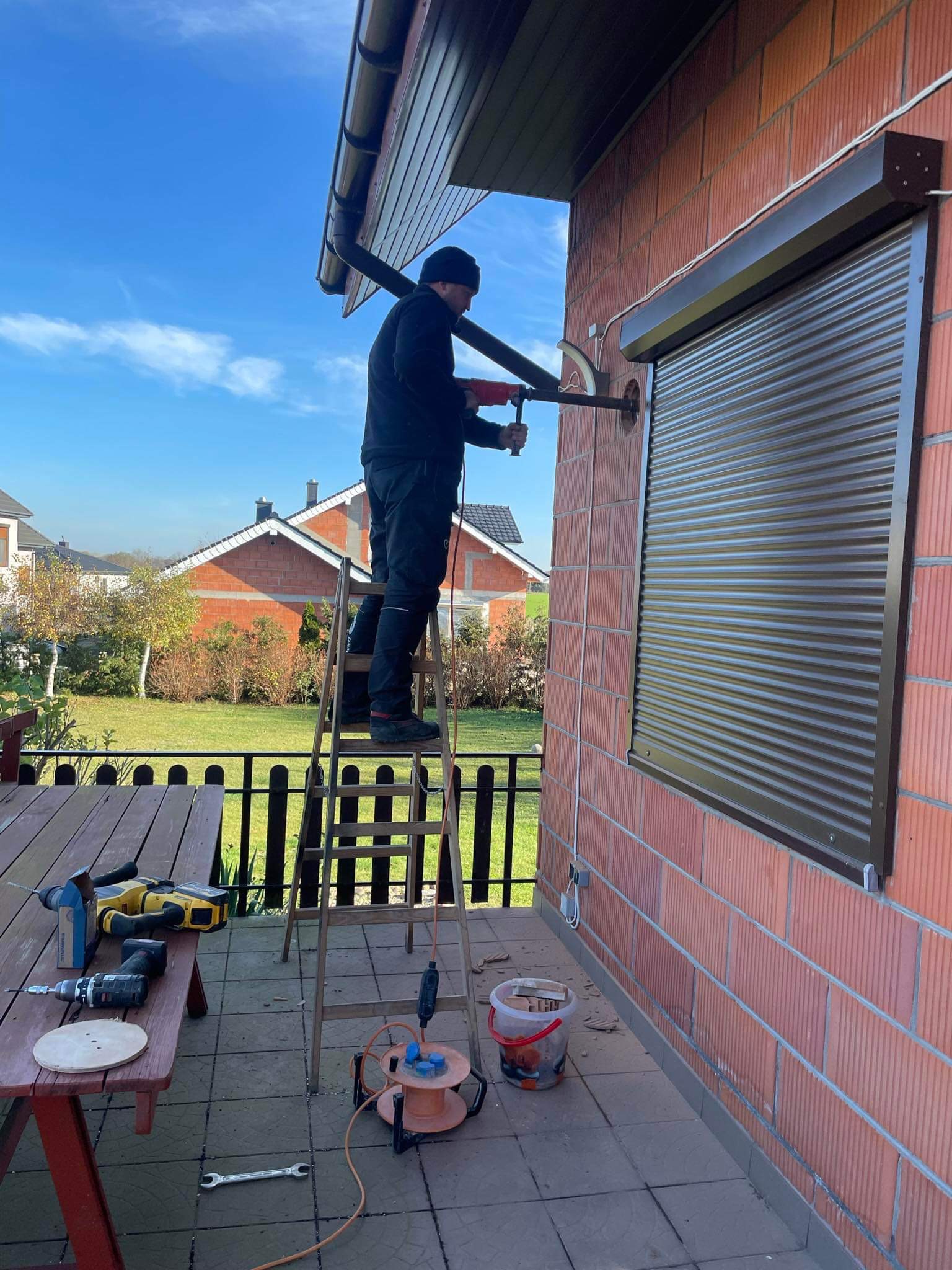 Monteraż rolety zewnętrznej na ceglanym budynku; mężczyzna na drabinie wierci otwór w ścianie, narzędzia leżą na stole na tarasie, widok na dom w tle.