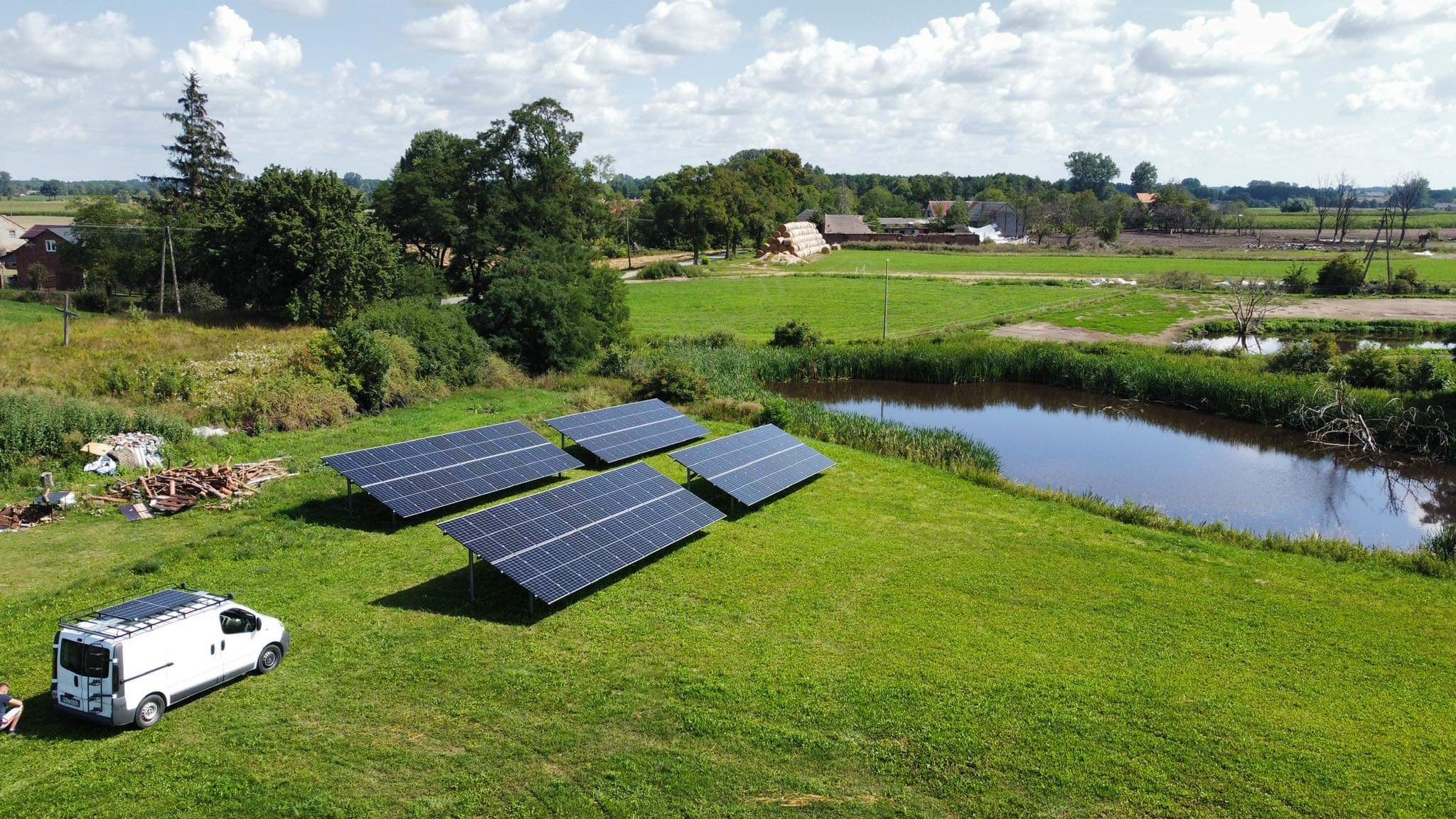 Cztery panele słoneczne na zielonej łące, obok stawu i z widokiem na wiejskie zabudowania, biały bus z panelami na dachu w lewym dolnym rogu, błękitne niebo z chmurami.