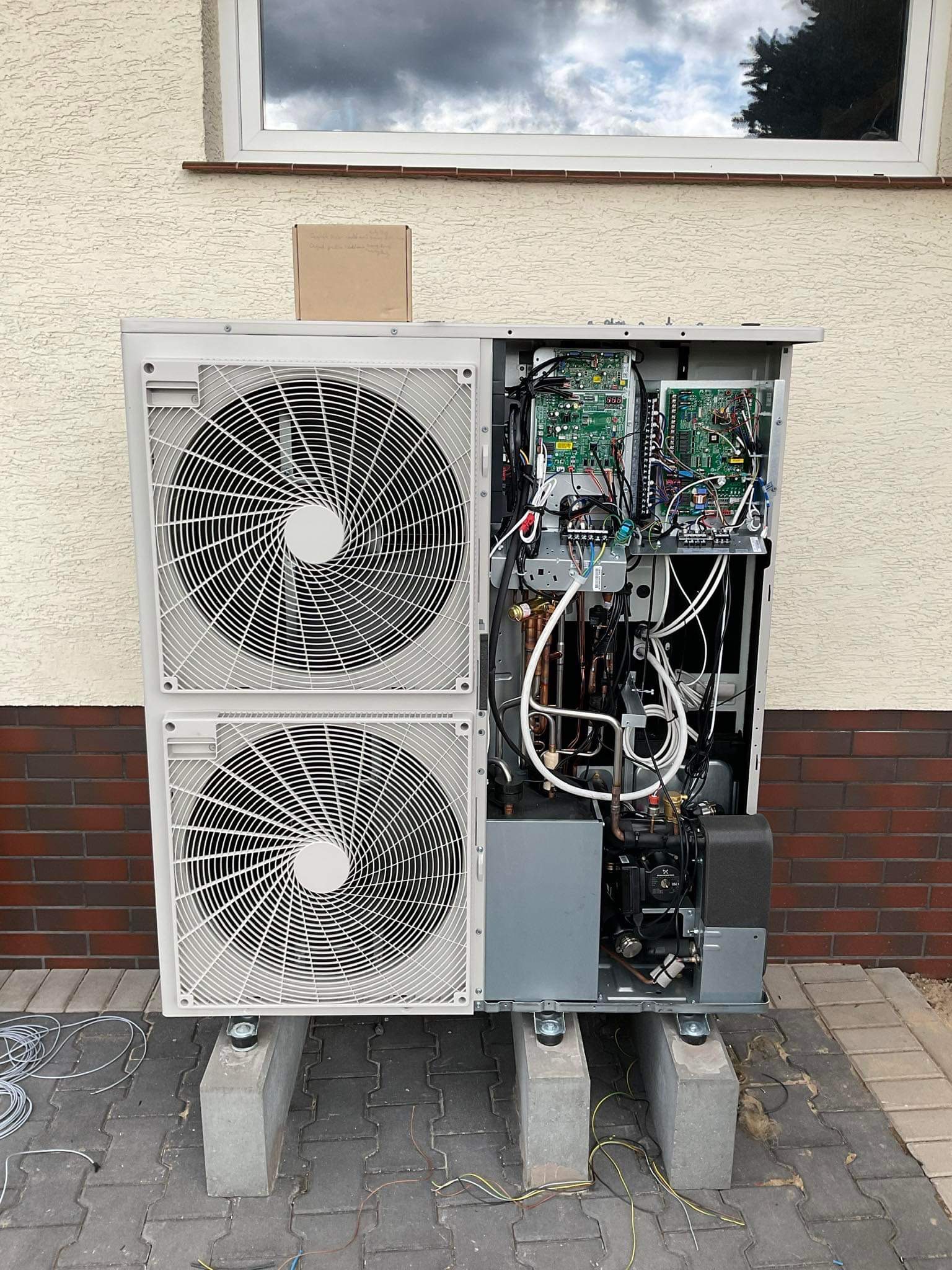 Otwarta obudowa pompy ciepła z widocznymi podzespołami elektronicznymi i hydraulicznymi, umieszczona na betonowych blokach w Trzebuchowie, na tle elewacji budynku z ceglaną wstawką.