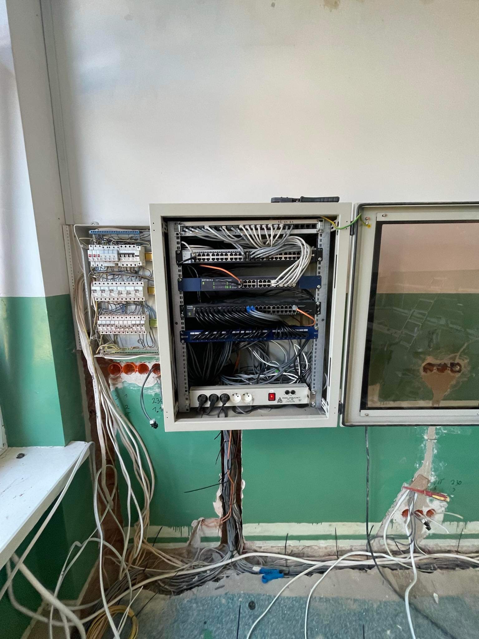 Otwarta szafa sterownicza z patch panelem, routerami i zasilaczem, obok skrzynka z bezpiecznikami; widoczne kable w ścianie i na podłodze, ściana w trakcie remontu.
