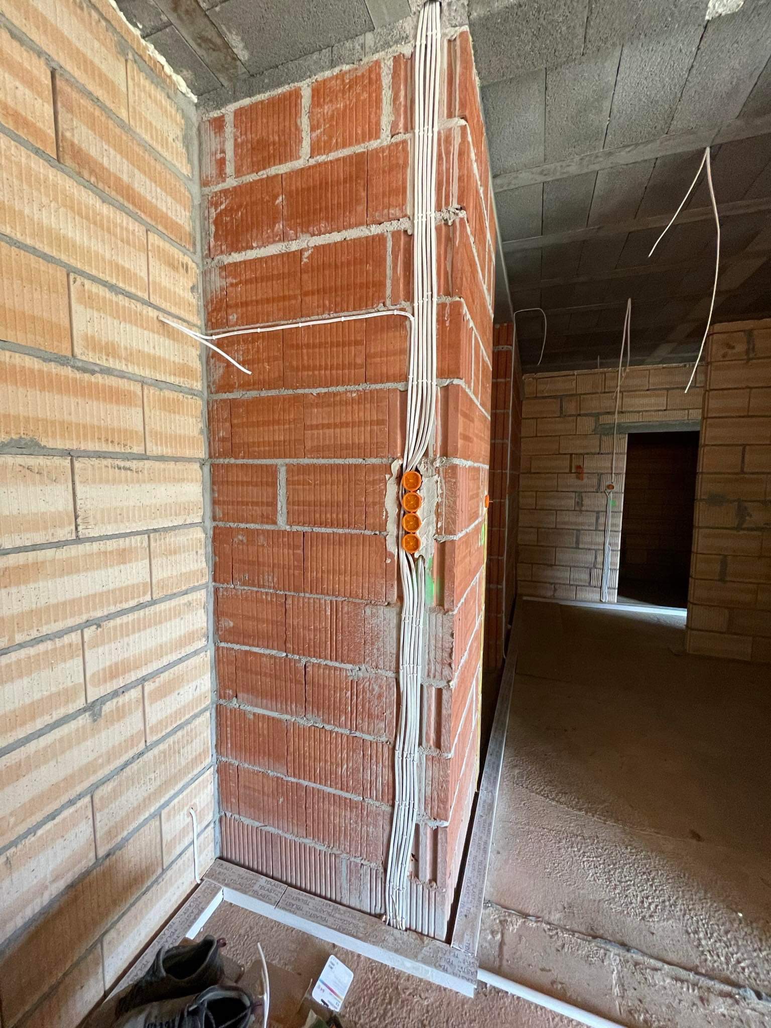 Instalacja elektryczna w surowym budynku: kable w białych peszlach prowadzone po ścianie z czerwonej cegły, widoczne pomarańczowe puszki instalacyjne, podłoga wylewka, w tle kolejne pomieszczenia.