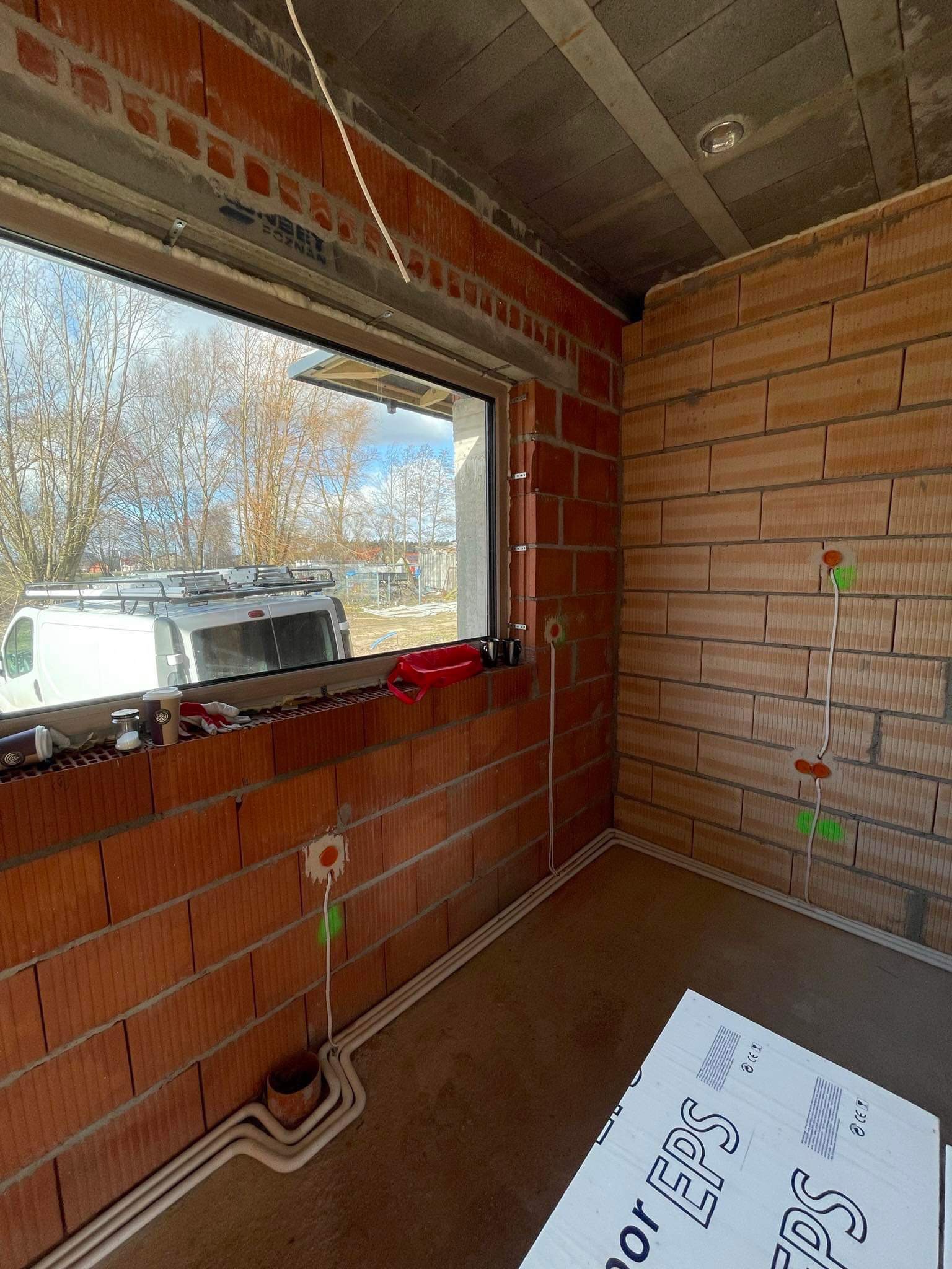 Wnętrze w trakcie budowy z widocznymi przewodami elektrycznymi poprowadzonymi po ścianach z czerwonej cegły, okno z widokiem na zewnątrz, kawa i akcesoria na parapecie, płyty EPS na podłodze.