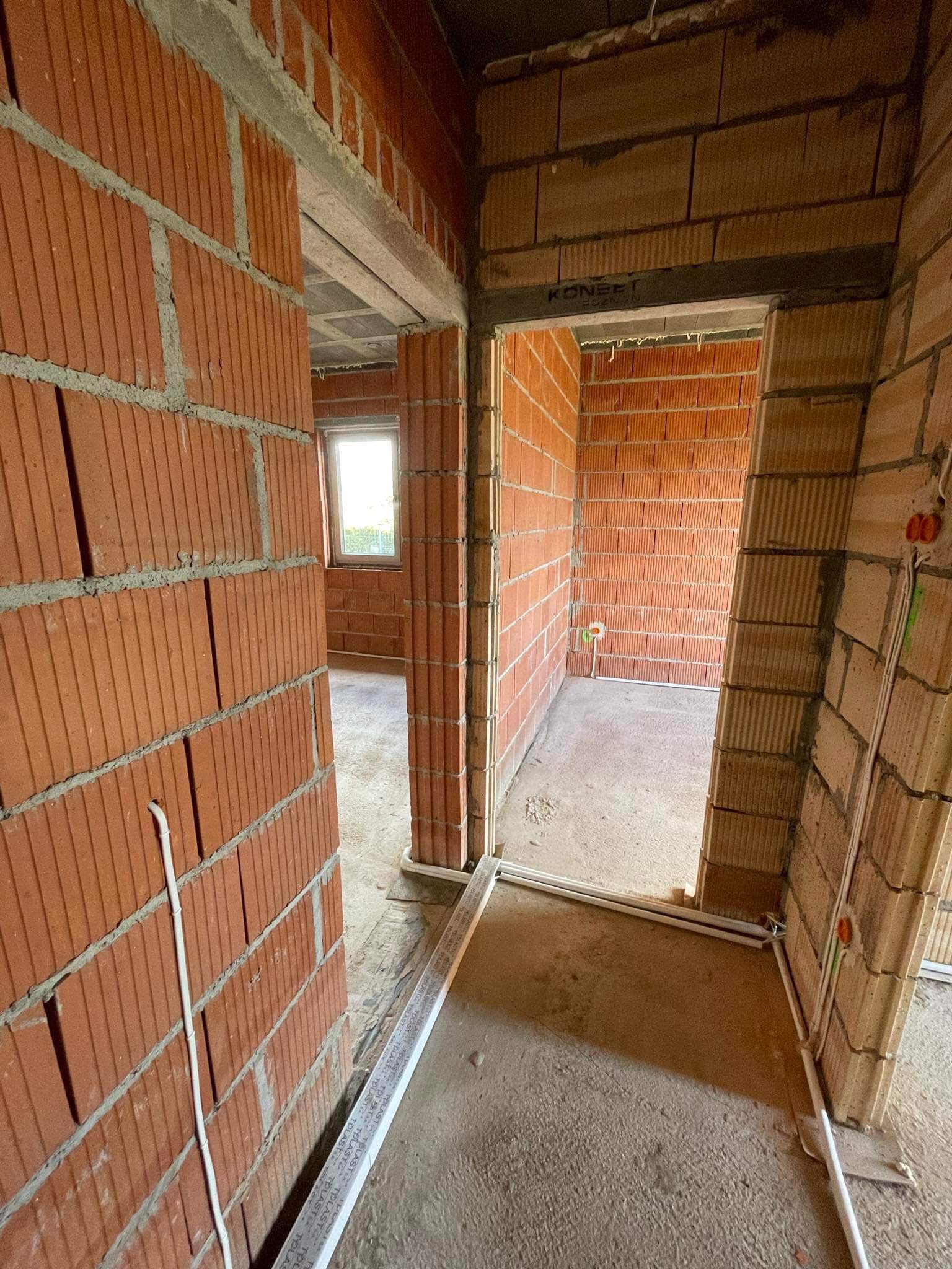 Surowy stan budynku w trakcie budowy, widoczne ściany z czerwonej cegły, betonowa podłoga i wystające przewody elektryczne, przygotowane pod instalacje.