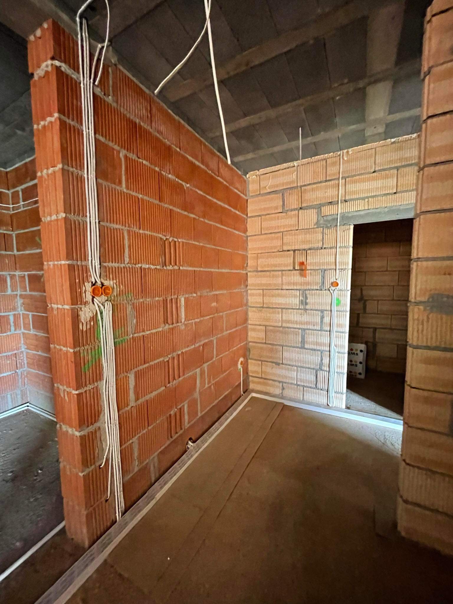 Wnętrze w trakcie budowy z widocznymi przewodami elektrycznymi prowadzonymi po ścianach z cegły ceramicznej, pomarańczowe puszki elektryczne zamontowane w ścianie, widoczny surowy strop betonowy.