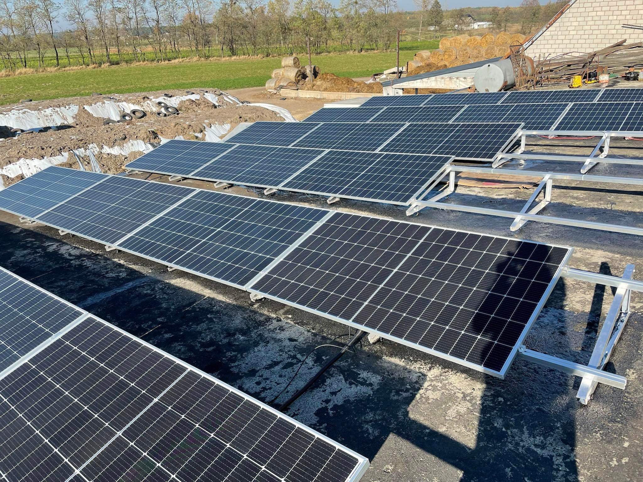 Panele słoneczne w trakcie montażu na dachu budynku gospodarczego, widoczne konstrukcje wsporcze, w tle pole i stogi siana.