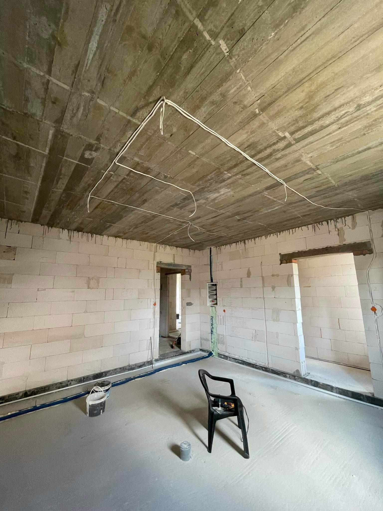 Surowy stan wnętrza budynku w trakcie prac: białe ściany z betonu komórkowego, betonowa wylewka podłogowa, widoczne przewody elektryczne pod sufitem, czarne krzesło na środku pomieszczenia...