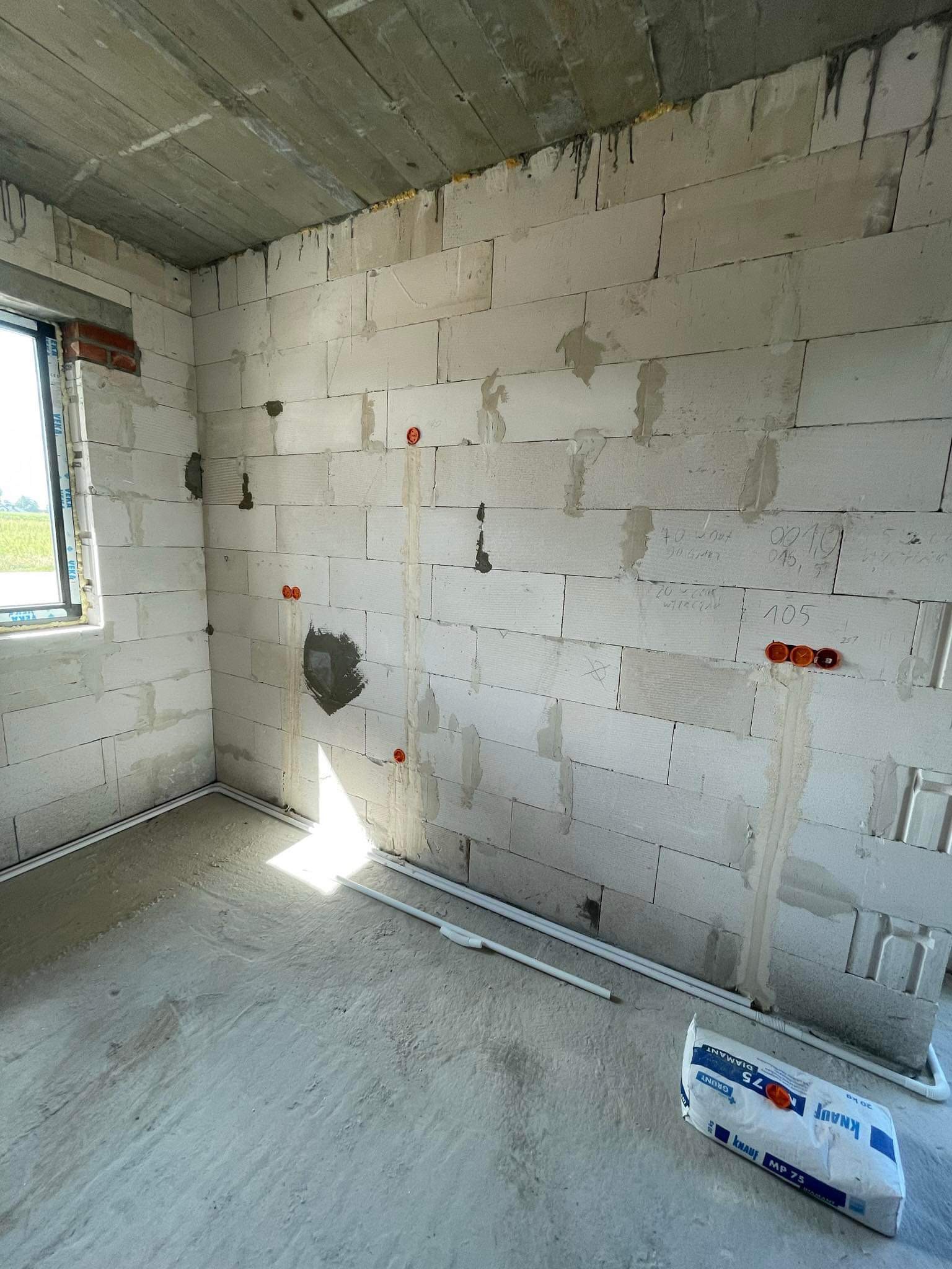 Surowy stan pomieszczenia z białymi bloczkami, widoczne przygotowane punkty elektryczne w ścianie, częściowo wylana wylewka na podłodze oraz leżący worek zaprawy Knauf MP 75.