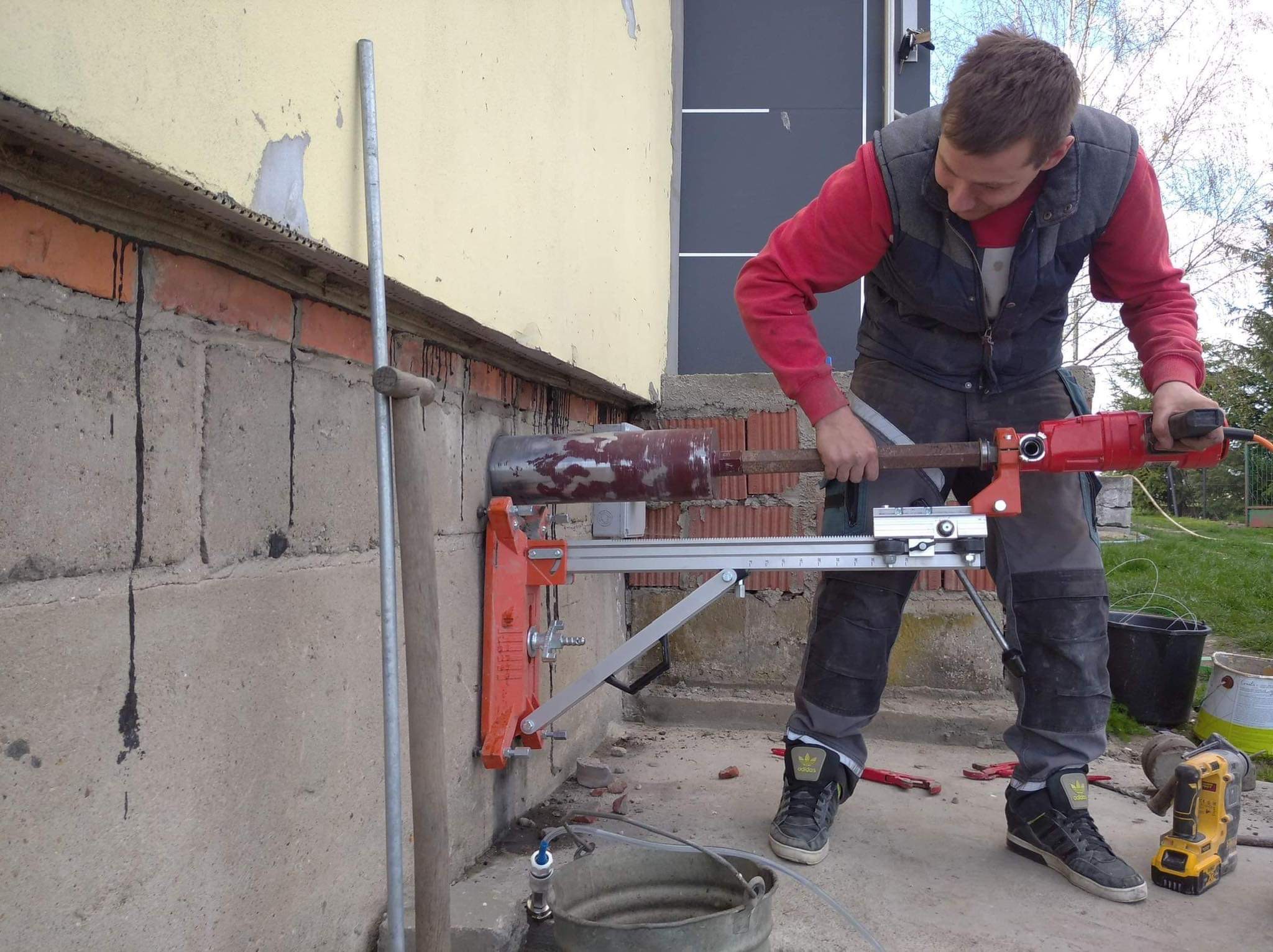 Pracownik wierci otwór w fundamencie budynku za pomocą wiertnicy diamentowej, widoczne narzędzia i osprzęt do wiercenia na mokro.