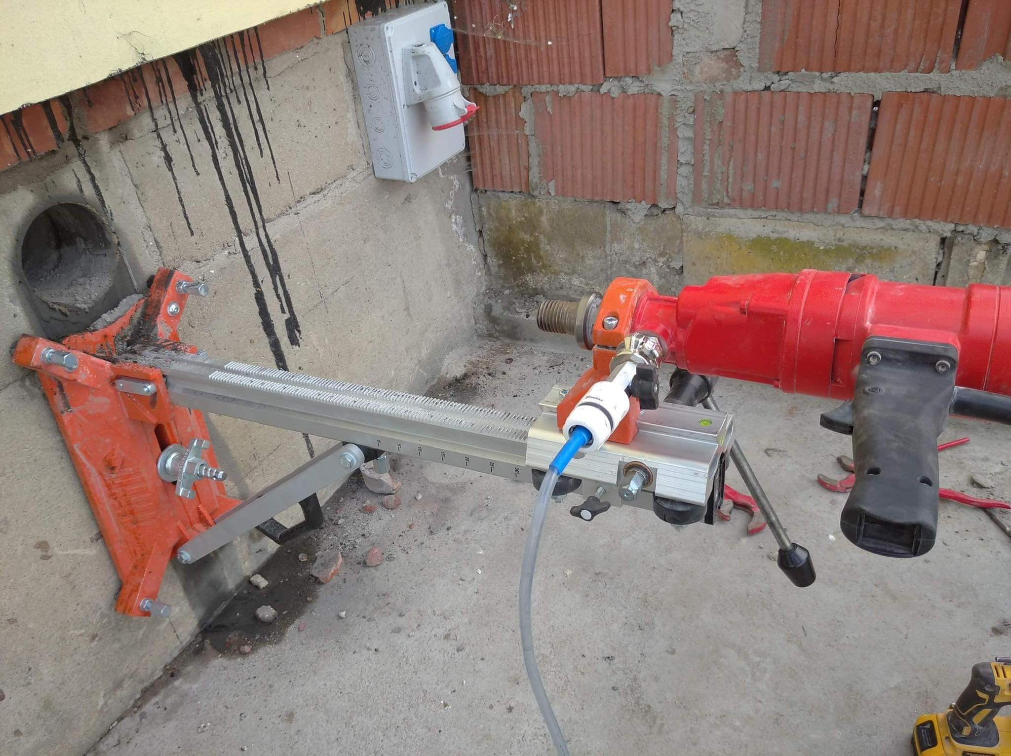 Czerwona wiertnica diamentowa przymocowana do ściany betonowej z widocznym otworem, przewodem doprowadzającym wodę i gniazdem elektrycznym w tle.