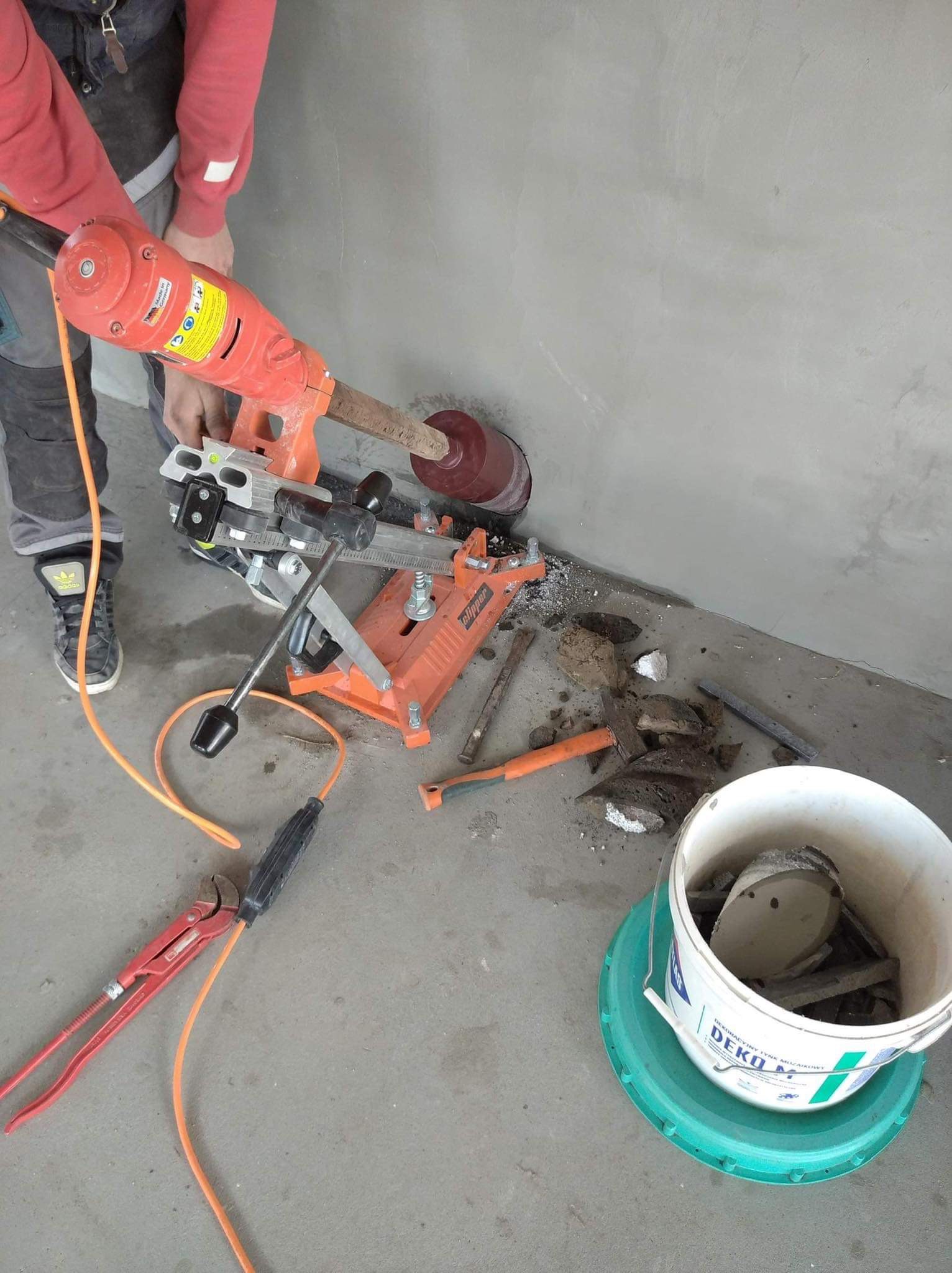 Wiercenie otworu w ścianie betonowej wiertnicą koronową z odsysaniem pyłu, widoczne odłamki betonu i wiadro na odpady, w tle fragment osoby obsługującej wiertnicę.
