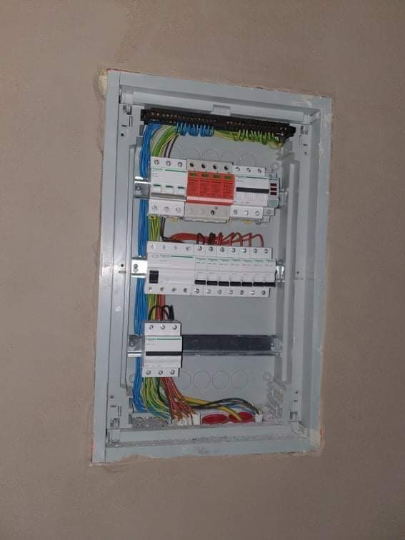 Otwarta skrzynka elektryczna z zainstalowanymi bezpiecznikami i różnokolorowymi przewodami na tle surowej ściany.