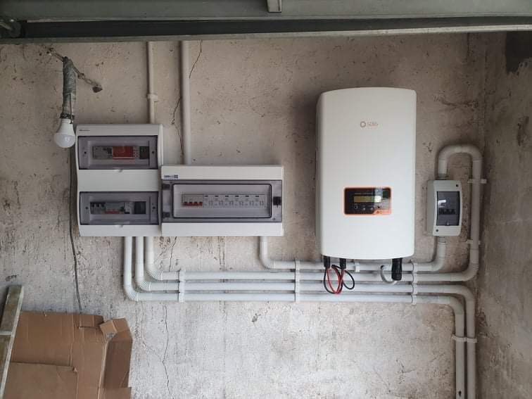 Zamontowany biały falownik z wyświetlaczem, połączony rurami PCV z dwoma rozdzielnicami elektrycznymi na surowej ścianie, oświetlony wiszącą żarówką na kablu.