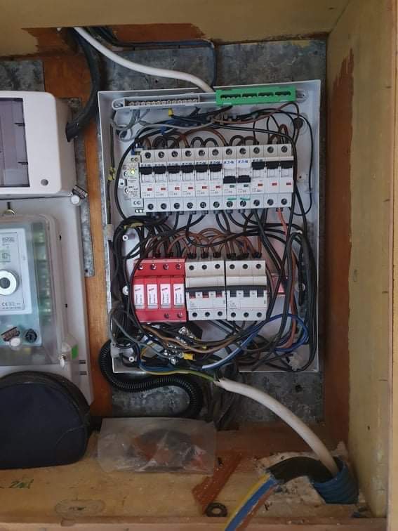 Otwarta skrzynka rozdzielcza z bezpiecznikami, ogranicznikami przepięć i przewodami, widoczne także licznik energii elektrycznej i fragment instalacji elektrycznej.