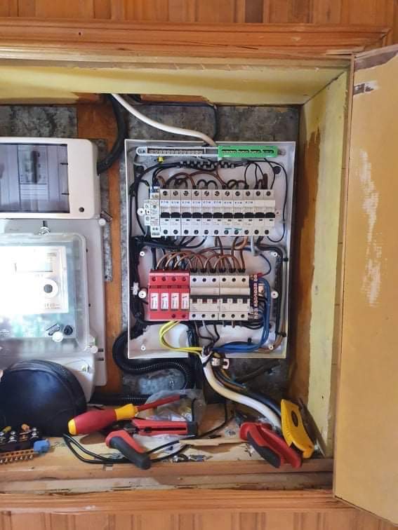 Otwarta skrzynka elektryczna z bezpiecznikami, przewodami i narzędziami, widoczny licznik energii i elementy instalacji elektrycznej w drewnianej obudowie.