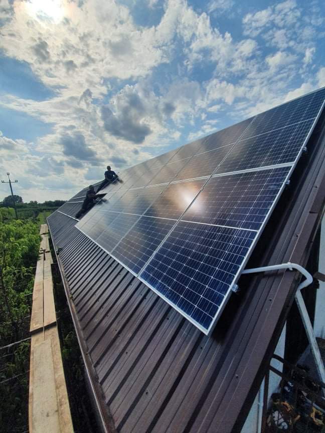 Montaż paneli słonecznych na brązowym dachu z blachy trapezowej, dwóch instalatorów w trakcie pracy, odbicie chmur w panelach, widok z dołu.