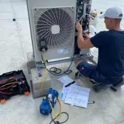 Serwisant w niebieskiej koszulce i czapce bejsbolowej, w kucki, podłącza przewody do klimatyzatora przemysłowego na dachu galerii handlowej; obok leżą narzędzia i dokumentacja techniczna.