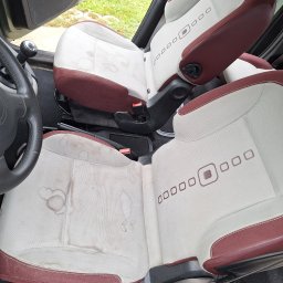 LAVENDA Ewa Samul - Tapicerka samochodowa z plamami na jasnym materiale, widoczne detale szwów i wzorów. Fotele wewnątrz pojazdu przed czyszczeniem, zarys kierownicy i dźwigni zmiany biegów.