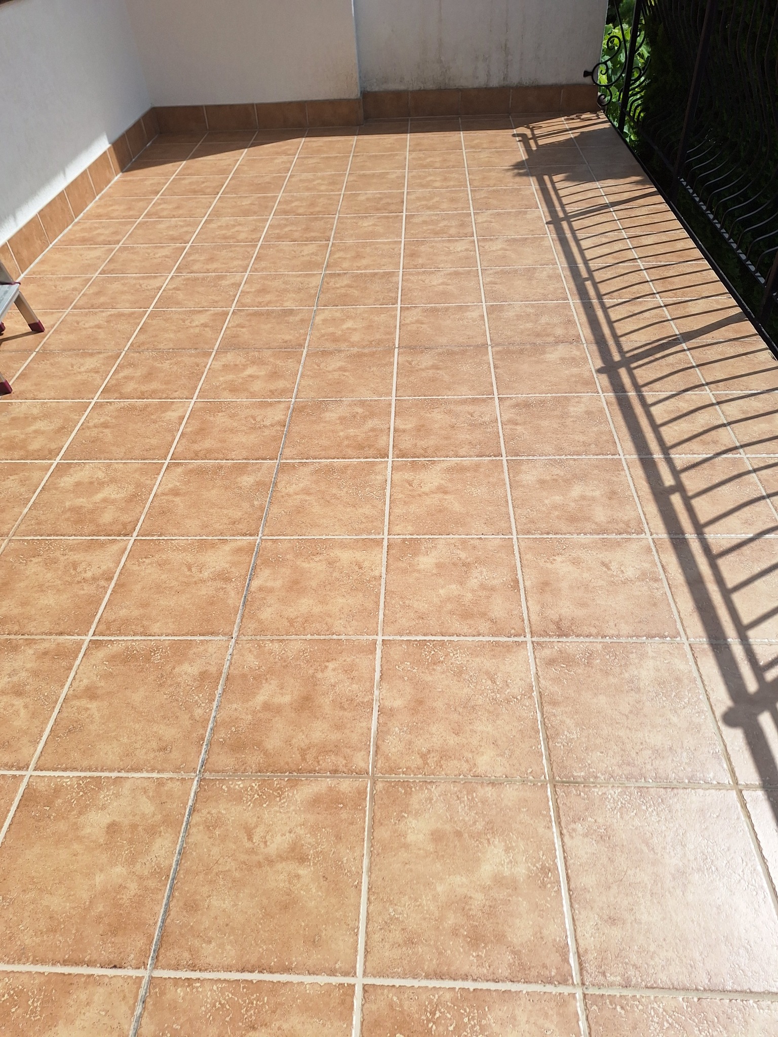 Czysty, brązowy taras wyłożony kwadratowymi płytkami ceramicznymi, z widocznym cieniem balustrady w słoneczny dzień.