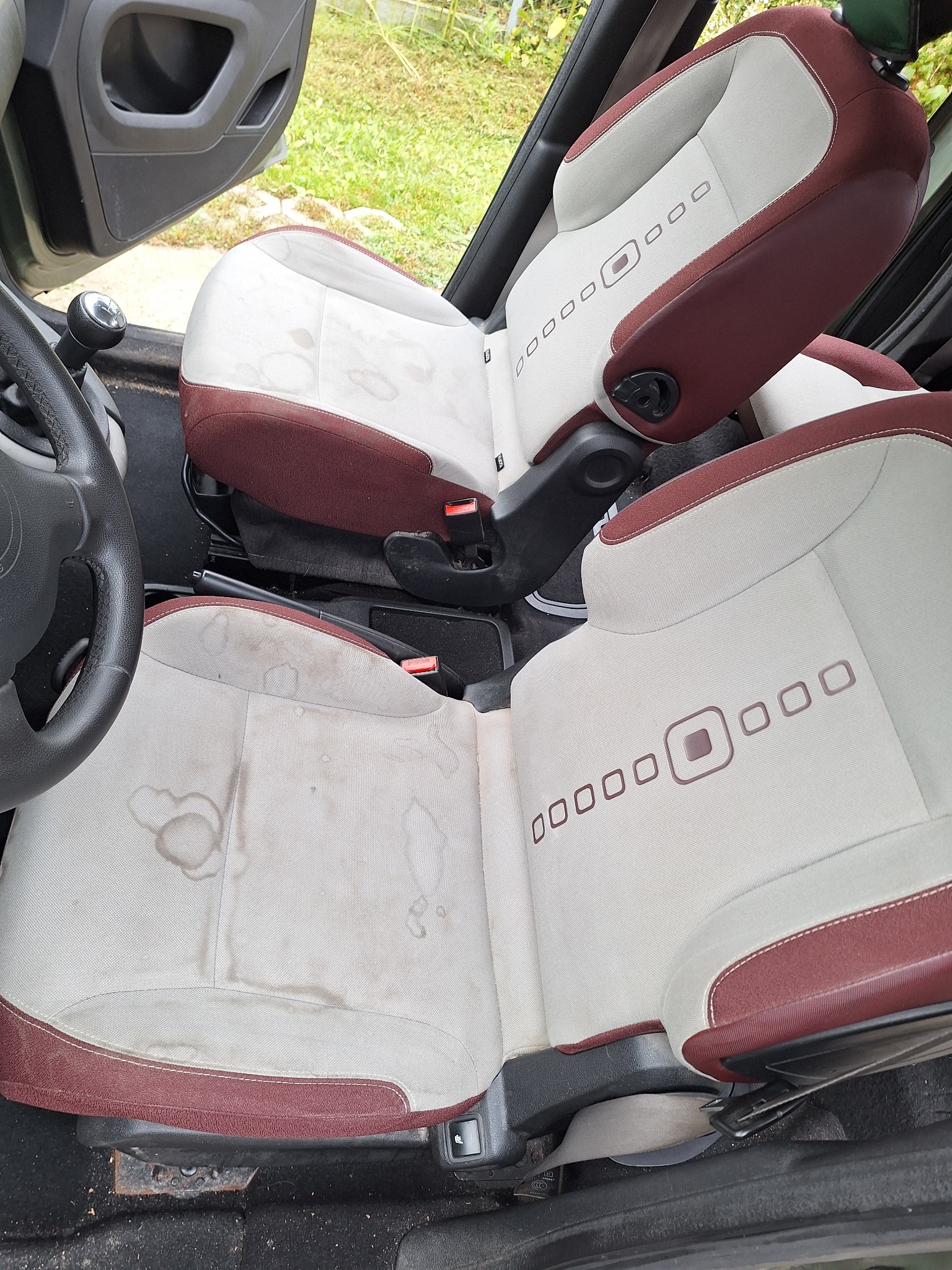 Tapicerka samochodowa z plamami na jasnym materiale, widoczne detale szwów i wzorów. Fotele wewnątrz pojazdu przed czyszczeniem, zarys kierownicy i dźwigni zmiany biegów.